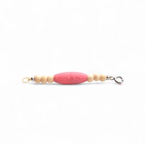 May include: A baby teething necklace with a pink, oval-shaped silicone bead in the center, flanked by small, light-colored wooden beads. The text "Kiddie King" is printed on the pink bead. The necklace has a clasp closure.