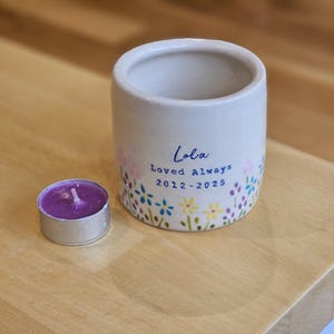 May include: A small, off-white ceramic candle holder with floral accents and the inscription "Lola Loved Always 2012-2025." A small, purple tea light candle sits beside it. The candle holder is cylindrical.