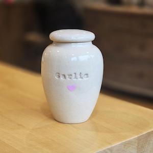May include: A cream-colored ceramic garlic keeper with a lid. The word "Garlic" is embossed on the front, below which is a small pink heart. The jar sits on a light-colored wooden surface.