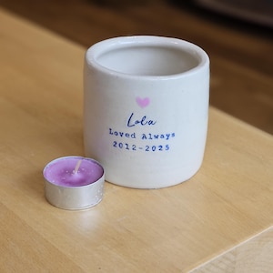 May include: A white ceramic candle holder with the name "Lola", the phrase "Loved Always", and the years 2012-2025 printed in blue. A small, round candle with a purple wax sits next to the holder.