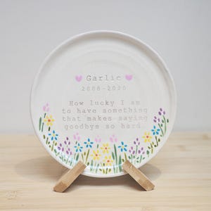 May include: A white ceramic plate with a floral design and the text "Garlic 2008-2020". The plate is supported by a wooden stand. The text reads "How lucky I am to have something that makes saying goodbye so hard."