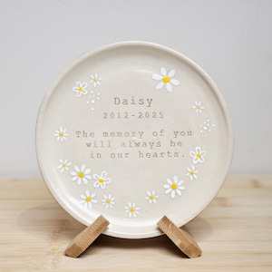 Puede incluir: Un plato de cerámica color crema con diseños de flores de margarita y el texto "Daisy 2012-2025 The memory of you will always be in our hearts." El plato se muestra sobre un soporte de madera.