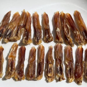 May include: A collection of dried duck feet, arranged on a white plate. The feet are a mix of light and dark brown, with a slightly translucent appearance. The texture appears to be chewy and dehydrated, suitable for pet treats.