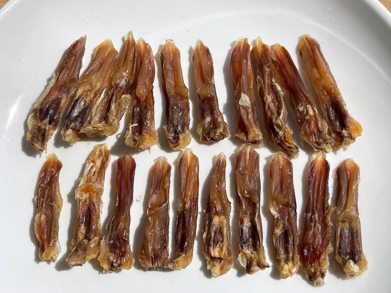 May include: A collection of dried duck feet, arranged on a white plate. The feet are a mix of light and dark brown, with a slightly translucent appearance. The texture appears to be chewy and dehydrated, suitable for pet treats.