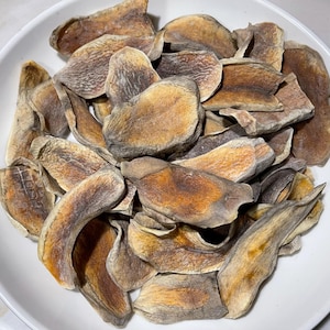 May include: A white bowl filled with dried, sliced fruit. The fruit pieces are various shapes and sizes, with a mix of brown, tan, and orange hues. The bowl is set against a light-colored surface.