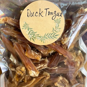 May include: A clear, sealed bag filled with dried duck tongues. A round, tan label with the words "Duck Tongue" in black script is affixed to the bag. The label is decorated with a green and white floral design.