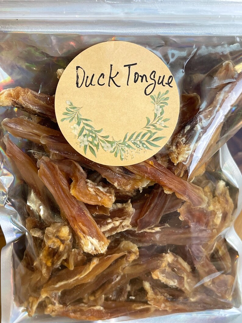May include: A clear, sealed bag filled with dried duck tongues. A round, tan label with the words "Duck Tongue" in black script is affixed to the bag. The label is decorated with a green and white floral design.