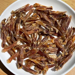 May include: A white plate filled with numerous pieces of dried, brown dog treats. The treats have a long, thin shape with a slightly curled appearance. The treats are a mix of light and dark brown colors.