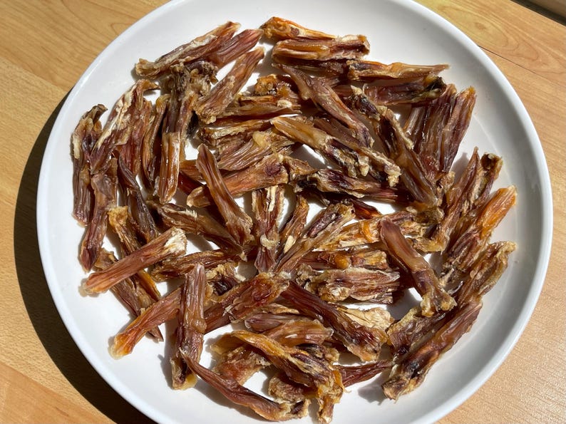 May include: A white plate filled with numerous pieces of dried, brown dog treats. The treats have a long, thin shape with a slightly curled appearance. The treats are a mix of light and dark brown colors.