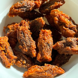 May include: A close-up shot of a white bowl filled with fried food. The food is a deep orange-brown color, with a crispy, textured exterior. The pieces are irregularly shaped and appear to be piled on top of each other.