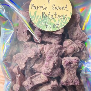 May include: A clear, iridescent bag filled with purple, bone-shaped treats. A round label reads "Beef & Purple Sweet Potatoes" with "4. oz" below. The treats are a light purple colour and appear to be dehydrated.