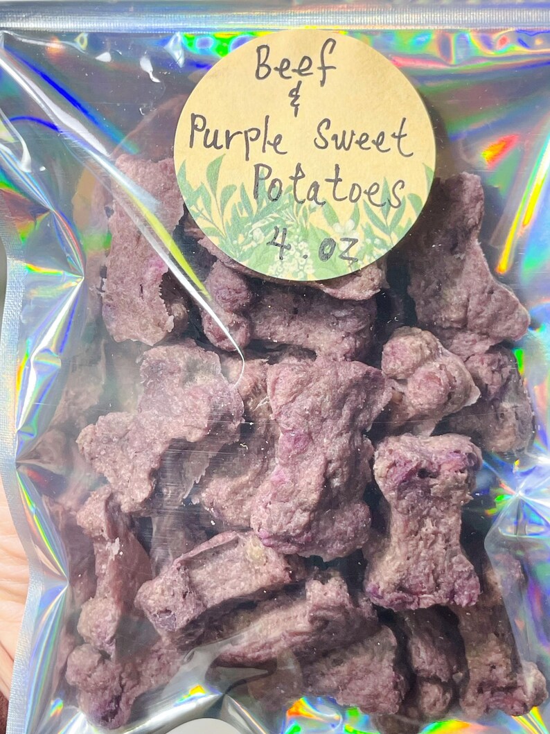 May include: A clear, iridescent bag filled with purple, bone-shaped treats. A round label reads "Beef & Purple Sweet Potatoes" with "4. oz" below. The treats are a light purple colour and appear to be dehydrated.