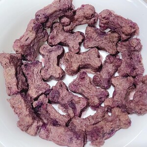 May include: A white bowl filled with dog treats shaped like bones. The treats are a mix of purple and brown, with a textured appearance. The treats are arranged in a pile, filling the bowl.