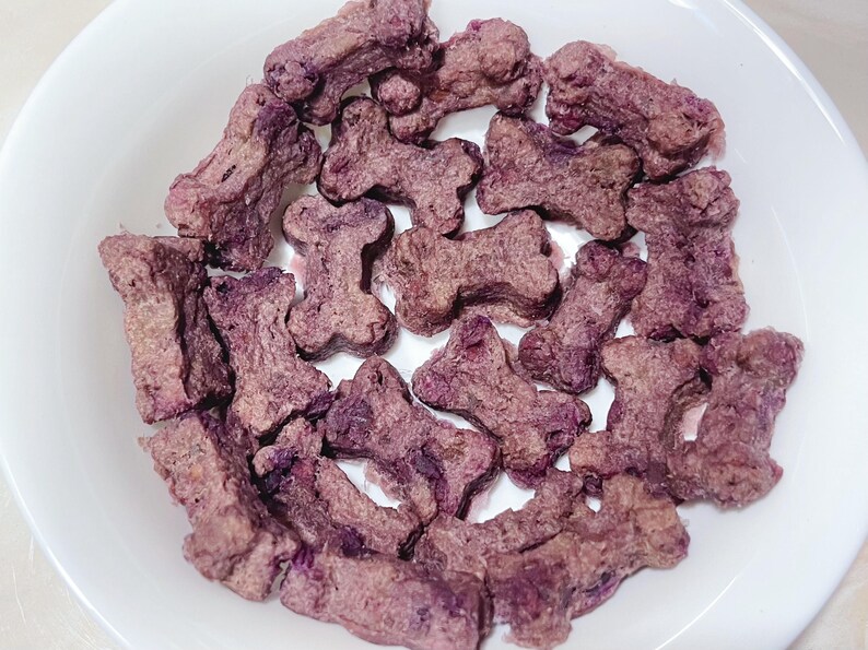 May include: A white bowl filled with dog treats shaped like bones. The treats are a mix of purple and brown, with a textured appearance. The treats are arranged in a pile, filling the bowl.