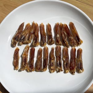 May include: A white plate holds a collection of dried, brown duck feet. The feet are arranged in two rows, showcasing their natural shape and texture. The plate sits on a light wooden surface.