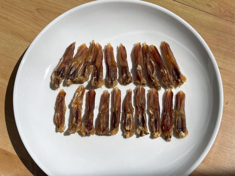 May include: A white plate holds a collection of dried, brown duck feet. The feet are arranged in two rows, showcasing their natural shape and texture. The plate sits on a light wooden surface.