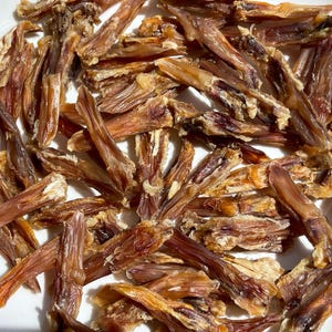 May include: A close-up shot of a white plate filled with dried, brown, and tan pet treats. The treats are long, thin, and irregularly shaped, with a slightly translucent appearance. The lighting highlights the texture and color variations.