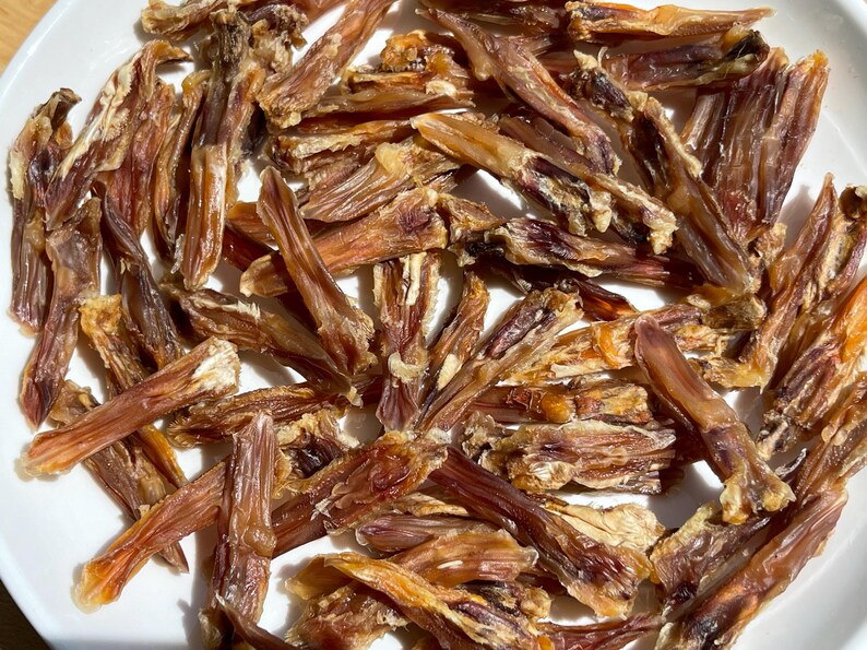 May include: A close-up shot of a white plate filled with dried, brown, and tan pet treats. The treats are long, thin, and irregularly shaped, with a slightly translucent appearance. The lighting highlights the texture and color variations.