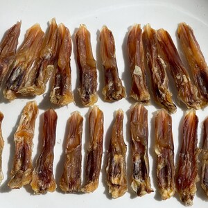 May include: A collection of dried, brown dog treats arranged on a white plate. The treats have a textured appearance and vary in size, with some showing darker brown areas. The lighting highlights the details of the treats.