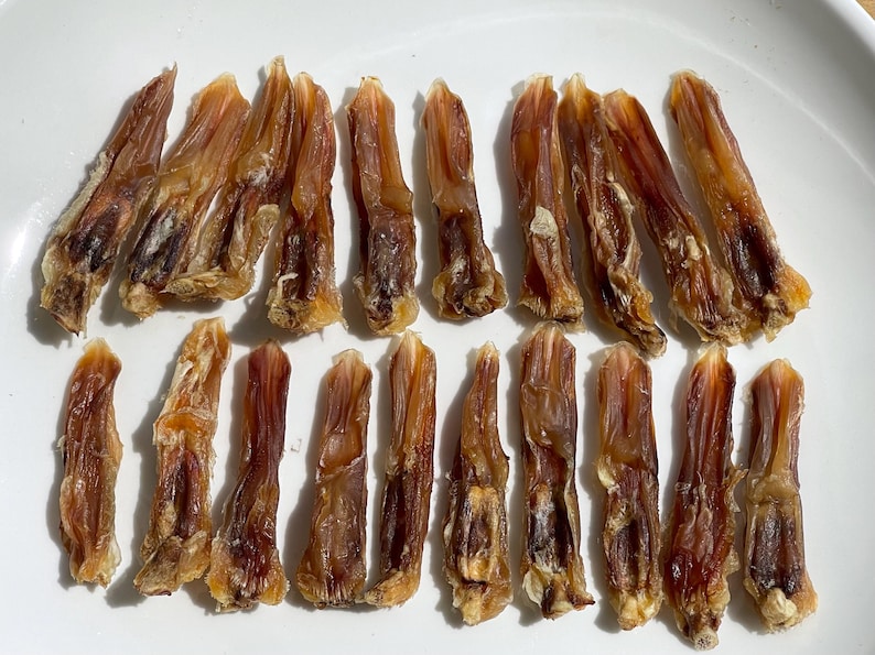 May include: A collection of dried, brown dog treats arranged on a white plate. The treats have a textured appearance and vary in size, with some showing darker brown areas. The lighting highlights the details of the treats.