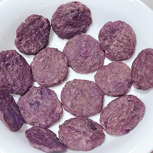 May include: A white bowl filled with a dozen round, purple-colored food items. The food items have a textured surface and appear to be a type of baked good or snack. The bowl is set against a light background.