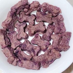 May include: A white bowl containing purple dog biscuits in the shape of bones. The treats are various shades of purple and brown, with a textured surface. The bowl sits on a light surface.