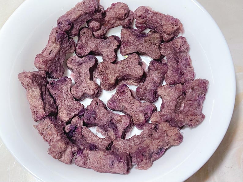 May include: A white bowl containing purple dog biscuits in the shape of bones. The treats are various shades of purple and brown, with a textured surface. The bowl sits on a light surface.