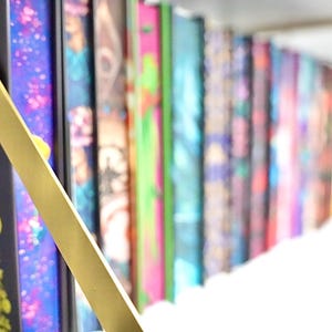 May include: A selection of books with vibrant, patterned covers. The spines showcase diverse designs, including floral, abstract, and geometric patterns. A gold-coloured object is in the foreground.