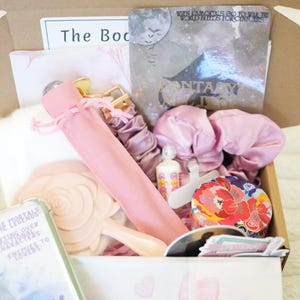May include: A cardboard box containing a selection of pink and white items. These include a hand mirror, scrunchies, a small bottle, and a decorative tape roll. There is also a book and a card with the text "Fantasy Day".