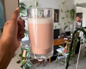 Peut inclure: Une tasse en verre transparent remplie d'une boisson brun rosé, surmontée d'une couche de mousse blanche. La tasse, avec une anse, est tenue par une personne. L'arrière-plan est une pièce avec des plantes et un bureau.
