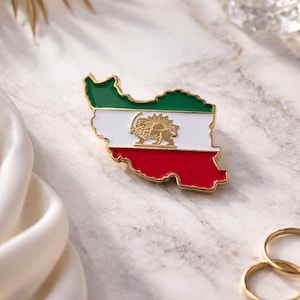 May include: A gold-edged pin in the shape of Iran, featuring the country's flag colors: green, white, and red. The white section displays a golden lion and sun emblem. The pin is set against a marble surface.