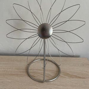 Puede incluir: Escultura de flor de metal plateado con base circular y esfera central. Los pétalos de la flor son de alambre fino, creando un diseño abierto. La escultura se exhibe sobre una superficie de madera clara.