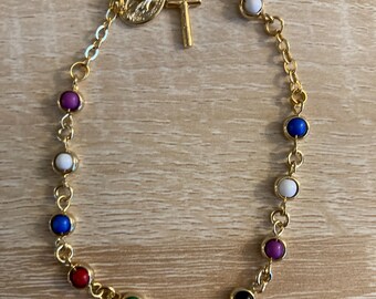 Puede incluir: Pulsera dorada con una serie de cuentas redondas y coloridas. La pulsera incluye un pequeño dije de cruz dorada y un medallón. Las cuentas son de varios colores, incluyendo azul, rojo, verde y rosa.