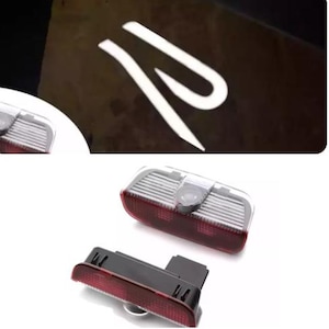 May include: Car door projector lights. The image shows a white light projection of a stylized letter "R" on a dark surface. The image also shows the projector lights themselves, which are rectangular with a red lens and a black base.