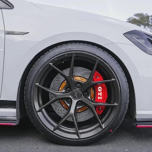 以下が含まれることがあります： 黒いリムとタイヤが付いた車のホイールのクローズアップ。ブレーキキャリパーは赤で、白で「GTI」と書かれています。ブレーキローターは金色です。車は白です。