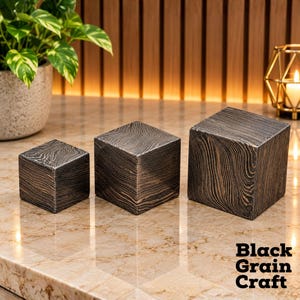 Pode incluir: Três cubos de madeira de tamanhos variados dispostos em fila, cada um com um acabamento de madeira escura carbonizada com padrões de veios pretos e castanhos. O texto Black Grain Craft está impresso a preto no canto inferior direito.