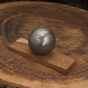 May include: A patterned metal sphere, approximately 2 inches in diameter, rests on a small, rectangular piece of wood. The sphere has a swirling, layered design. The wood is a warm brown color, set against a textured wooden background.