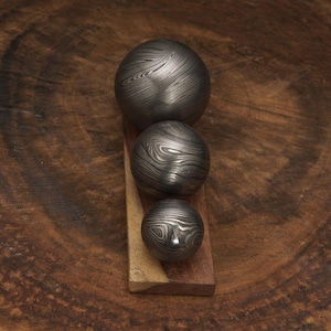 May include: Three decorative metal spheres with a unique swirling pattern, arranged vertically on a wooden base. The spheres vary in size, with the largest at the top and the smallest at the bottom. The metal has a dark, textured appearance.