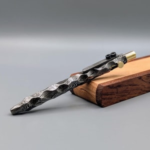 May include: A textured metal pen with a unique wave-like pattern. The pen has a brass tip and a clip with two small screws. It rests on a wooden box, showcasing its craftsmanship and design. The pen's body has a dark, patterned finish.
