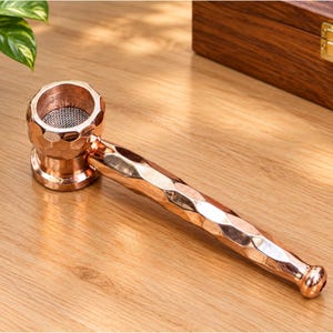 Pode incluir: Um cachimbo de metal na cor ouro rosa com uma textura martelada e facetada no fornilho e na haste. O fornilho circular apresenta uma malha metálica fina na base, e a haste é longa e cilíndrica com uma extremidade arredondada.