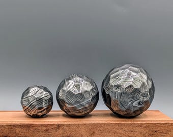 May include: Three textured metal spheres of varying sizes rest on a wooden base. The spheres have a patterned surface, with a dark, metallic appearance. The wooden base is rectangular and has a reddish-brown color. The background is a solid gray.