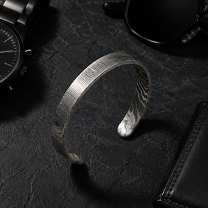 May include: A silver-toned metal cuff bracelet featuring a distinct, layered, wavy pattern across its entire surface. The bracelet has an open-ended design with rounded tips, showcasing a complex, wood-grain-like texture in shades of gray and silver.