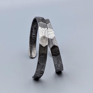 May include: Two silver cuff bracelets with an open design. The bracelets display a distinctive pattern of swirling lines and shapes, giving a textured look. The design is visible on the inside and outside of the bracelets.