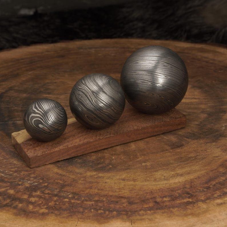 May include: Three metal spheres with a unique patterned surface rest on a small, rectangular piece of wood. The spheres vary in size, with the largest one on the right. The wood is a reddish-brown color, and the background is a dark, blurred texture.