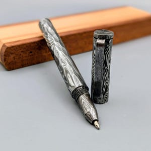 Puede incluir: Un bolígrafo de acero de Damasco con un patrón de vetas de madera. El bolígrafo tiene una punta plateada y un capuchón a juego. El bolígrafo está sobre una superficie gris con un portalápices de madera en el fondo.