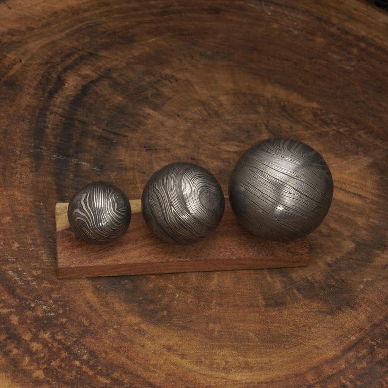 May include: Three metal spheres with a unique swirling pattern, displayed on a small wooden base. The spheres vary in size, with the largest one having a diameter of approximately 3 inches. The metal has a dark, textured appearance.