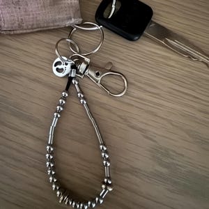 Può includere: Un portachiavi con perline argentate con un moschettone e un piccolo ciondolo con un design a impronta. Il portachiavi è attaccato a un portachiavi con una chiave dell'auto e un portachiavi in tessuto.