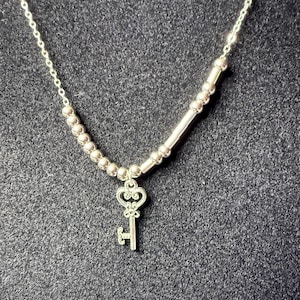May include: A silver-toned necklace with a key pendant. The necklace features a delicate chain and a series of small, round beads. The key pendant has an ornate design, adding a touch of elegance to the jewelry.