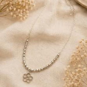 以下が含まれることがあります： 繊細なシルバーのネックレス、花のペンダント付き。ネックレスは、小さな丸いビーズと円筒形のバーが付いたチェーンが特徴です。ペンダントは詳細な花のデザインです。ネックレスはベージュの布の上にあり、背景にはドライフラワーがあります。