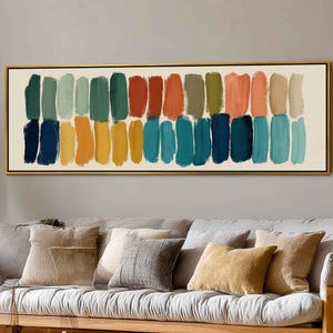 Puede incluir: Impresión artística abstracta con filas de pinceladas coloridas en tonos verdes, azules, naranjas y amarillos. La obra de arte está enmarcada en un marco dorado y cuelga sobre un sofá gris con cojines decorativos.
